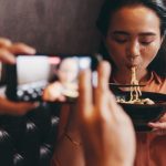 Mujer joven come un plato de fideos chinos mientra otro le graba con el móvil