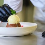 La mano de un chef con guante negro coloca el último detalle de un postre